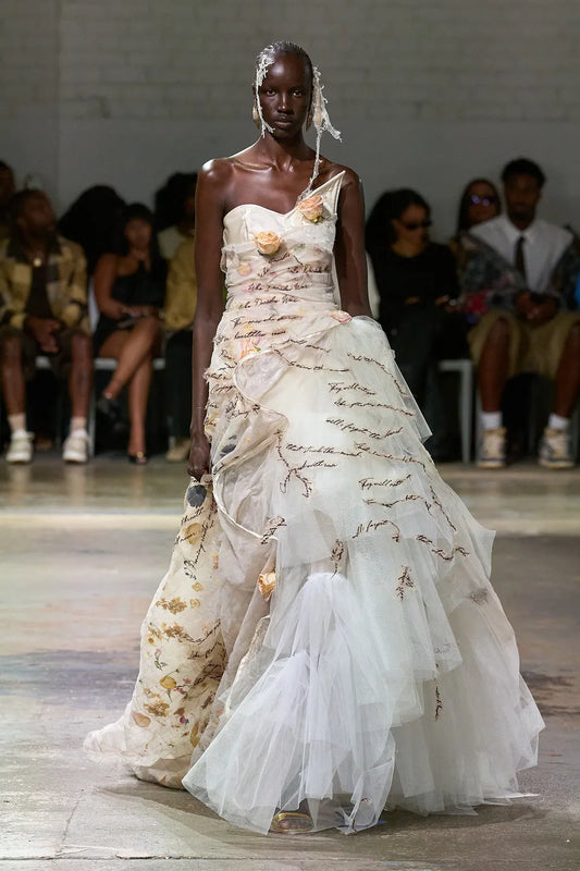 Model wearing a white, intricately designed dress on a runway with an audience.